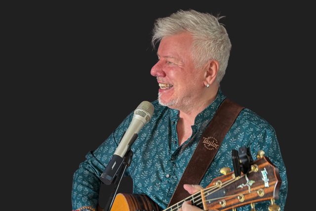 Musician Gordon Mark Webber smiling while playing acoustic guitar and singing during a live music event at Essence Restaurant in Tunbridge Wells