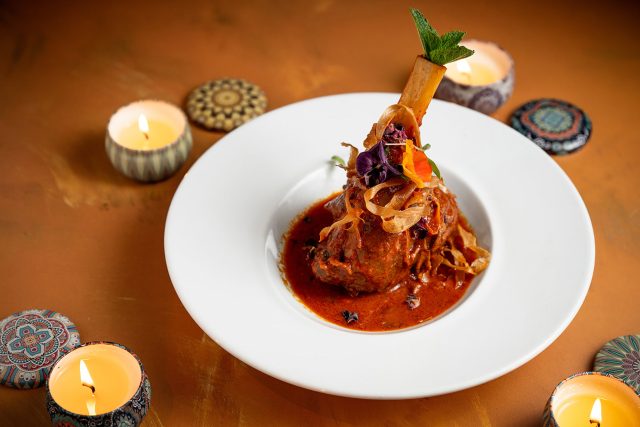 Spiced lamb shank curry garnished with edible flowers and mint, surrounded by glowing Diwali candles on a warm-toned table.