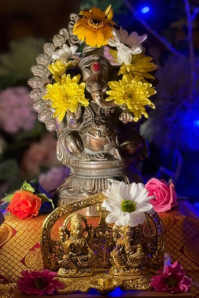 Bronze Lord Ganesha statue and golden Hindu deities decorated with fresh flowers for a Diwali celebration, illuminated by ambient blue lighting.
